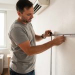Un homme mesure les surfaces sur un mur dans un salon lumineux afin de préparer l’installation d’une climatisation murale.