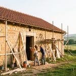 Ancienne maison de campagne en pierre en cours de rénovation, avec un échafaudage discret, trois artisans au travail et un paysage rural lumineux en arrière-plan.